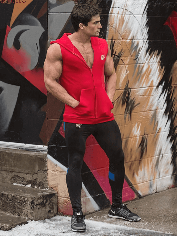 Man wearing a red sporty vest with zipper and hood, standing confidently by urban graffiti in leggings and sneakers.